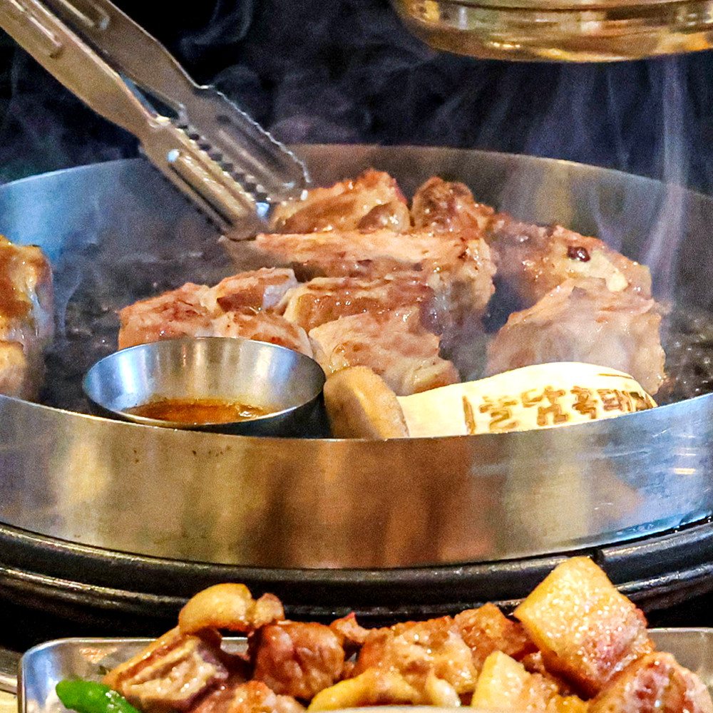 돌담흑돼지 연동 본점 — 불판에서 노릇하게 익어 가는 흑돼지와 집게, 가운데 멜젓 종지